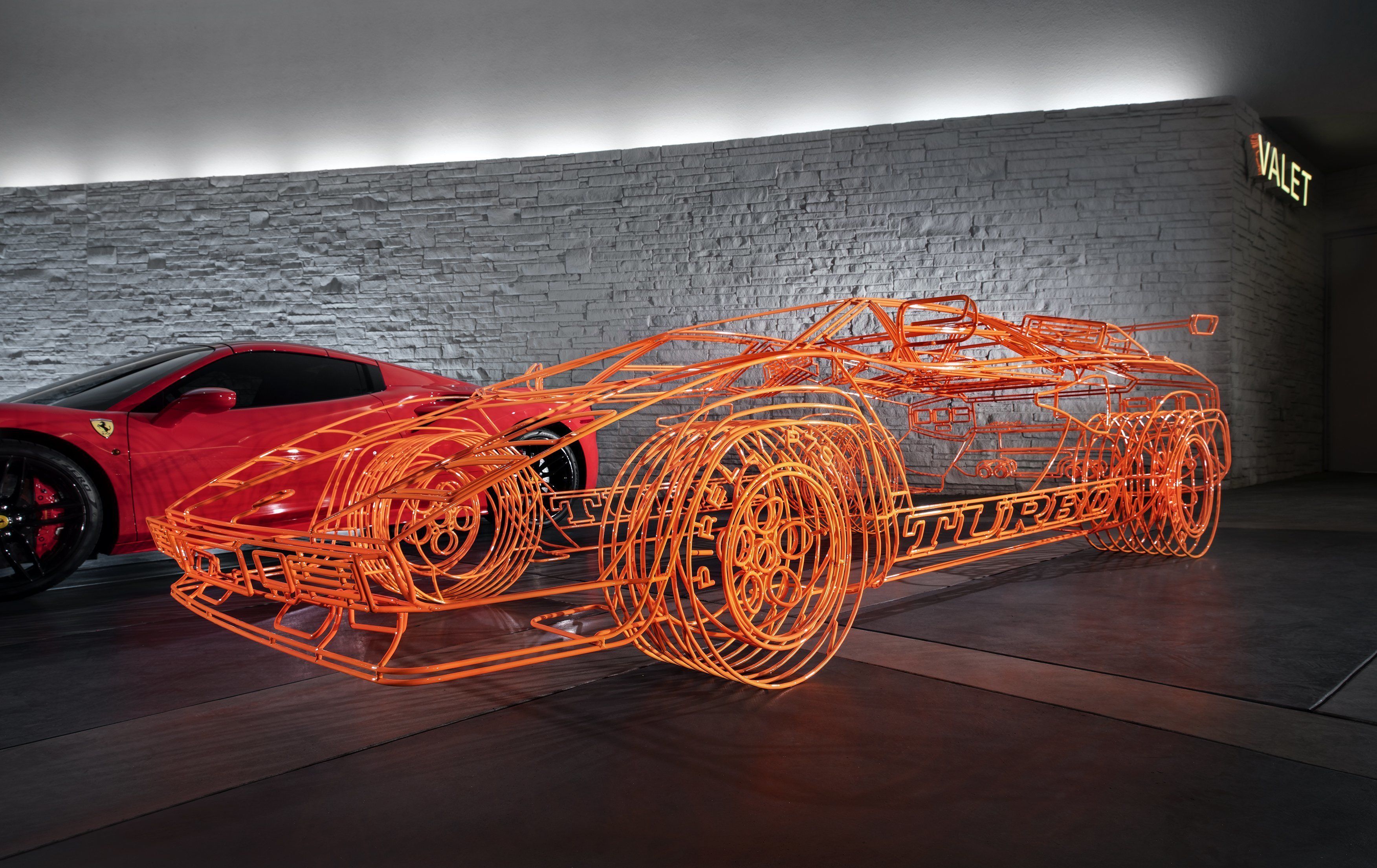 A red sports car with a transparent, orange wireframe structure in a room with a brick wall and a sign that reads "VALET".