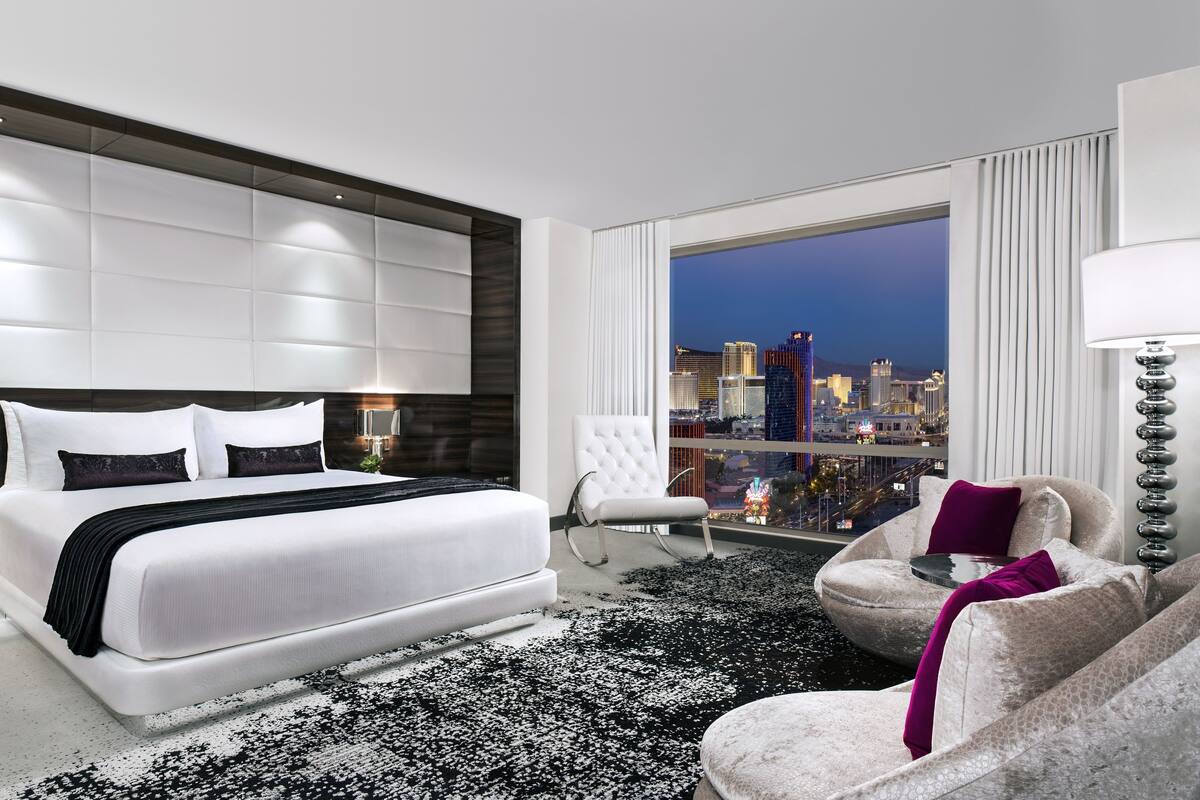 A modern hotel room with a large bed, a city view, a white armchair, and a small table with a lamp.