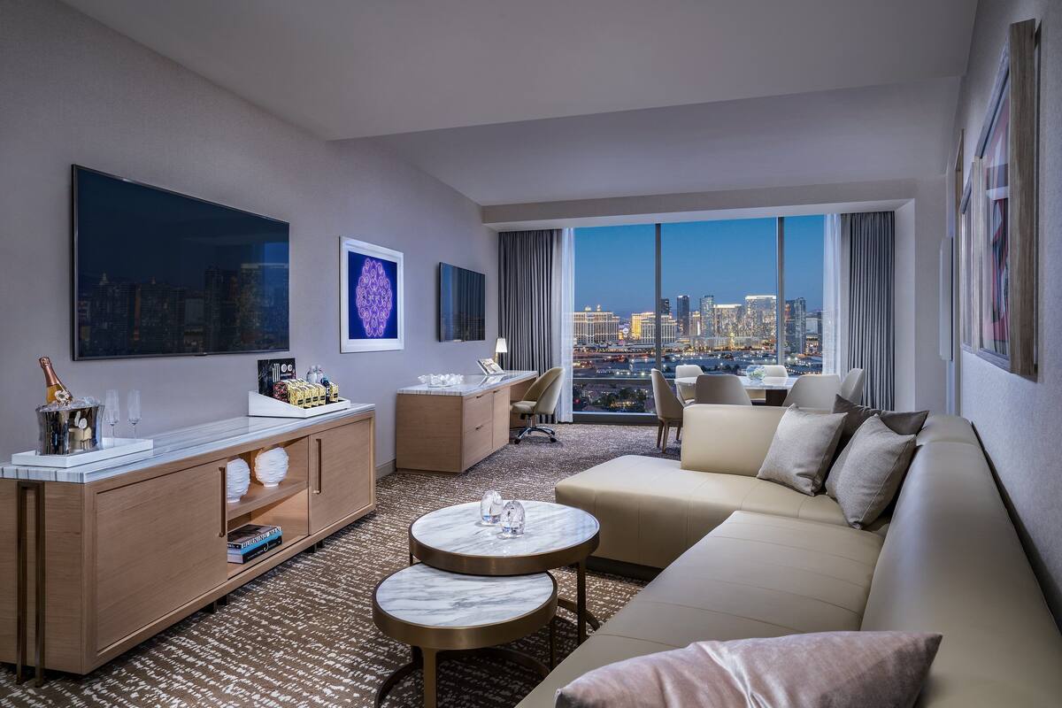 A modern hotel room with a large city view, a sofa, a dining table, and a TV.