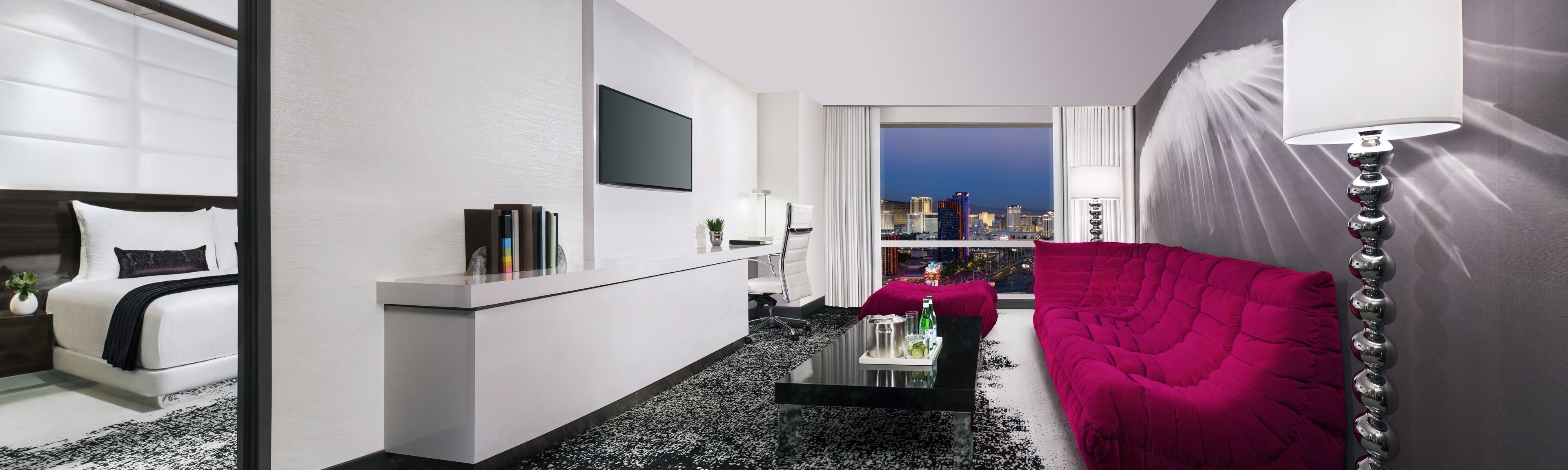 A modern hotel room with a white bed, a black and white patterned rug, a red sofa, a flat-screen TV, and a city view.