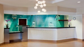 A reception area with a teal accent wall, a reception desk, a clock, and a hanging light fixture. - SpringHill Suites Los Angeles LAX/Manhattan Beach (Hawthorne)