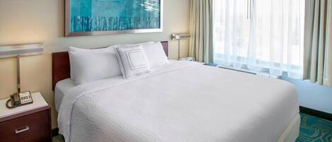 A hotel room with a large bed, a bedside table with a phone, a painting on the wall, and a window with curtains.