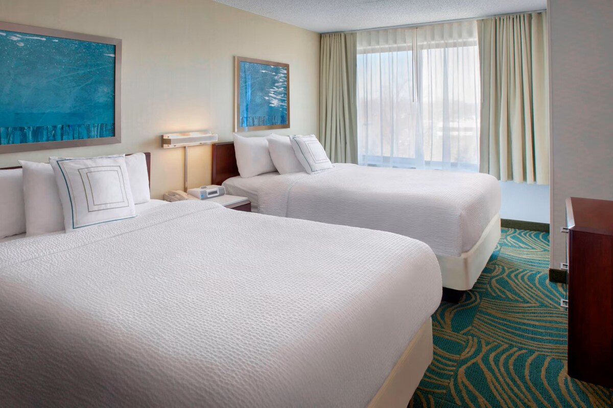 A hotel room with two beds, a nightstand, a lamp, and a large window with curtains.