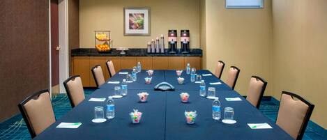 Un salón de conferencias con una larga mesa, sillas, botellas de agua y máquinas de café.