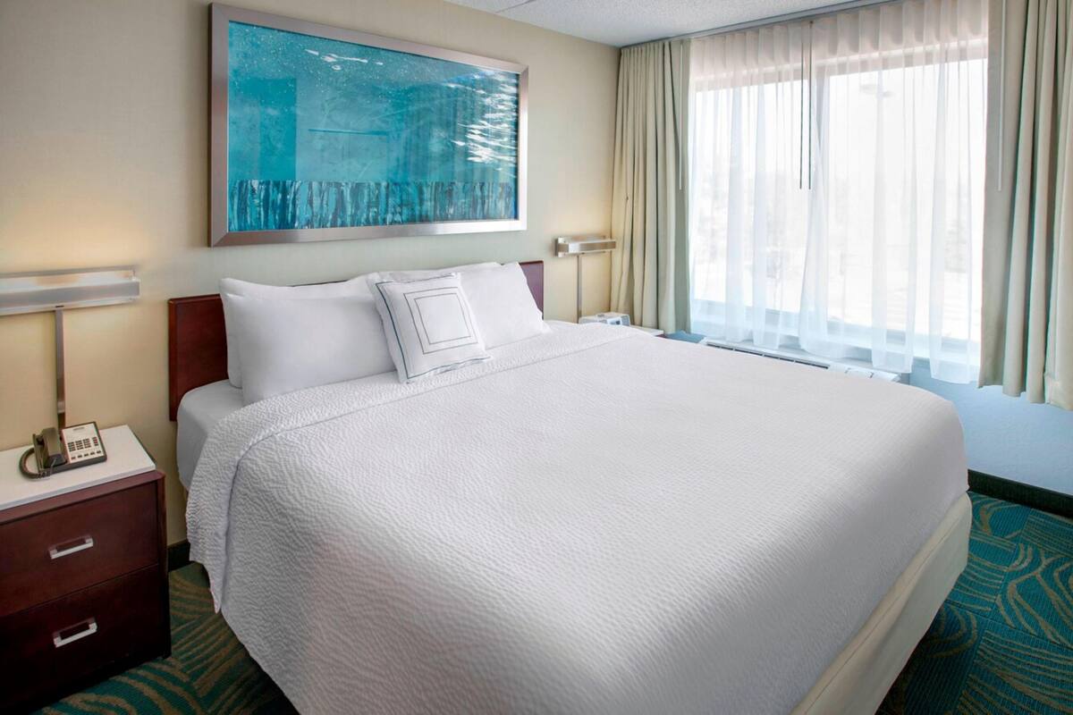 A hotel room with a large bed, a bedside table with a phone, a painting on the wall, and a window with curtains.