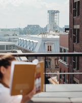 Una persona leyendo un libro en un balcón con vistas a un paisaje urbano con edificios y una torre.