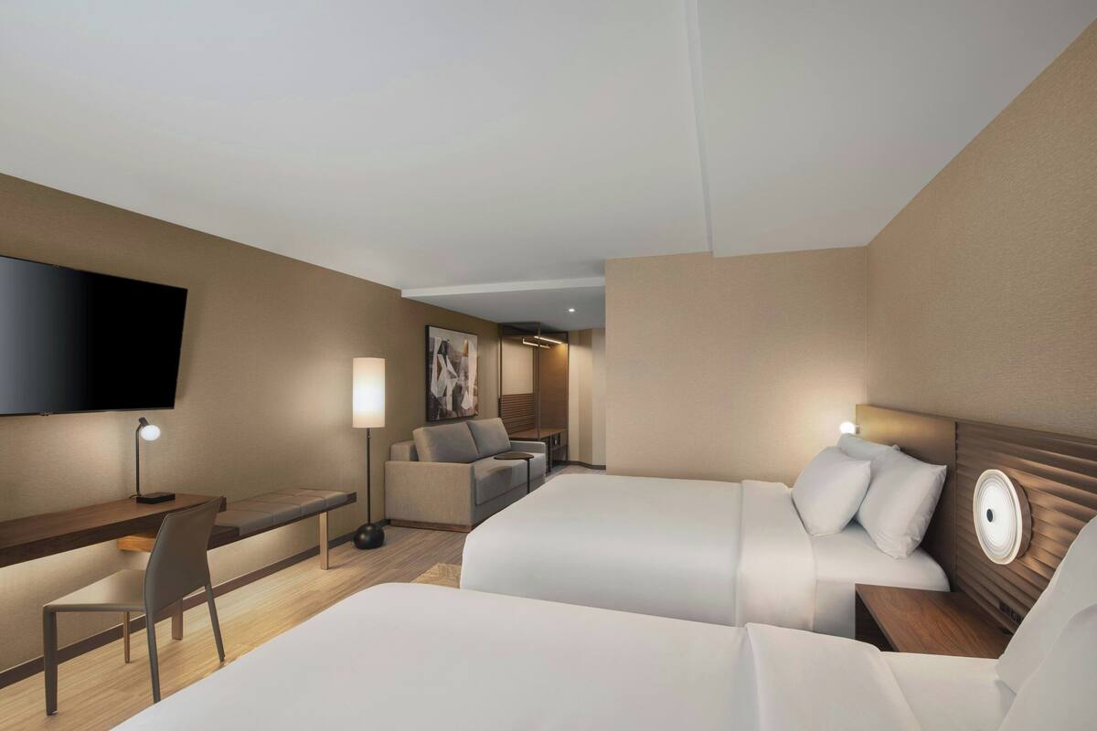 A hotel room with two beds, a desk, a chair, a flat-screen TV, and a wall-mounted artwork.