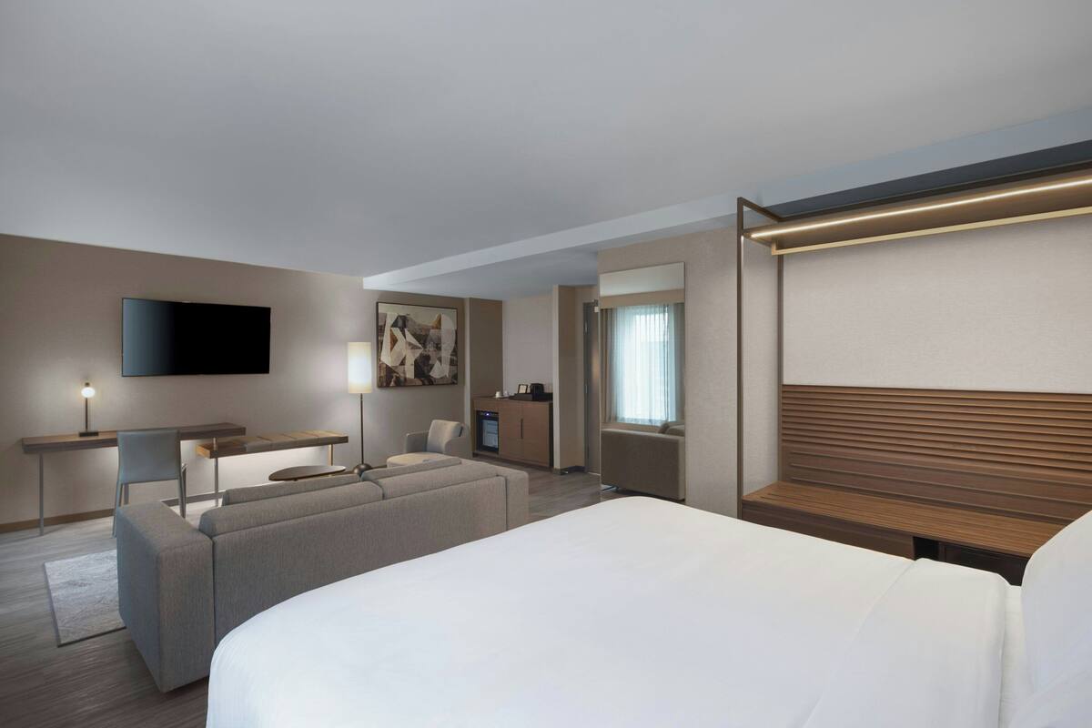 A modern hotel room with a large bed, a desk with a chair, a sofa, a TV mounted on the wall, and a window with curtains.