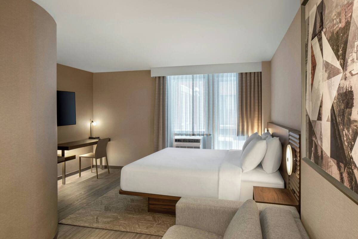 A modern hotel room with a large bed, a desk, and a chair. There is a television mounted on the wall and a window with curtains.