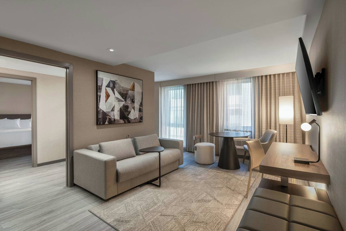 A modern hotel room with a sofa, a small table, a lamp, and a TV mounted on the wall.