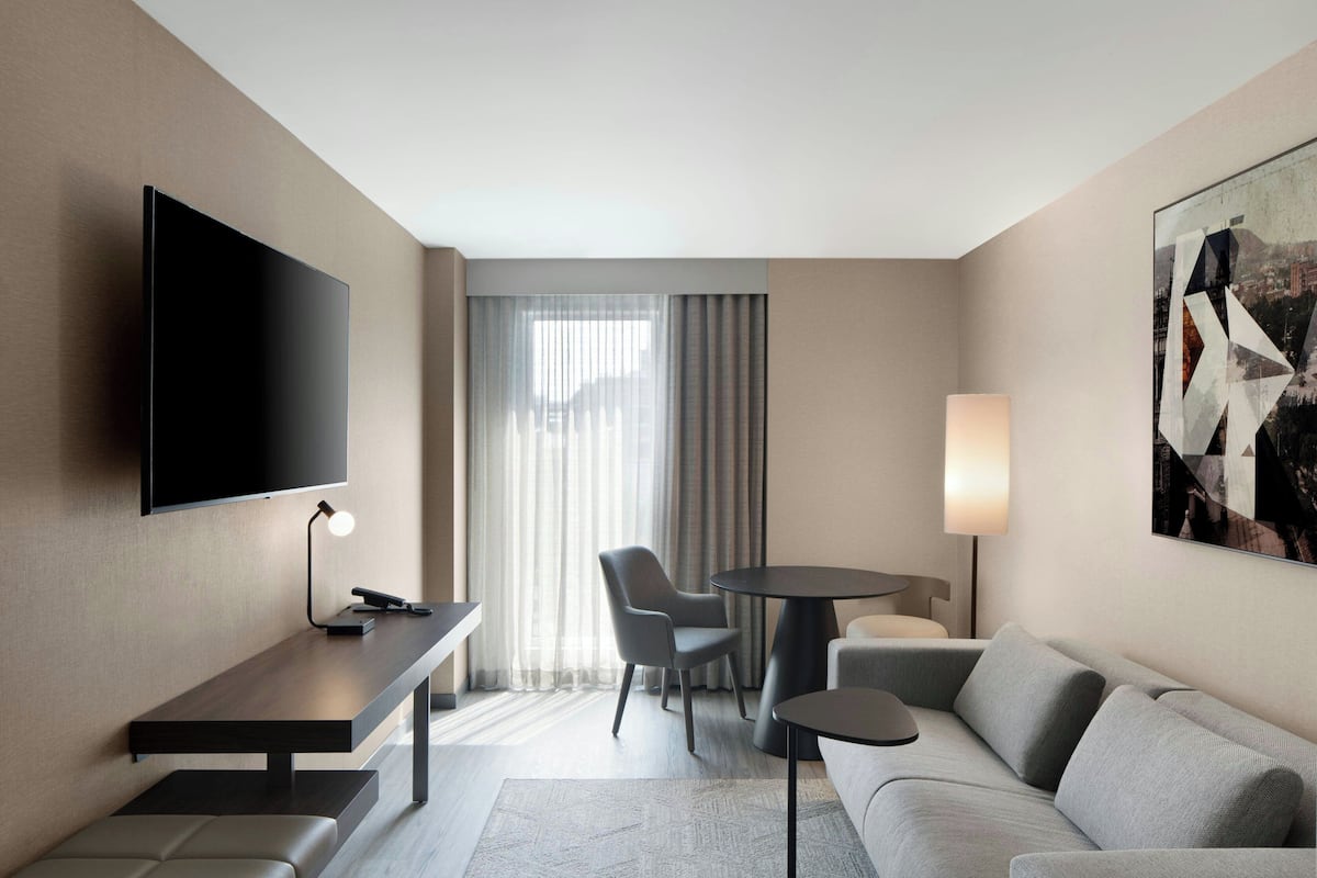 A modern hotel room with a flat-screen TV, a desk with a lamp, a sofa, a round table, and a chair.