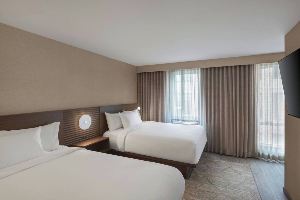 A hotel room with two beds, a flat-screen TV, and a large window with curtains.