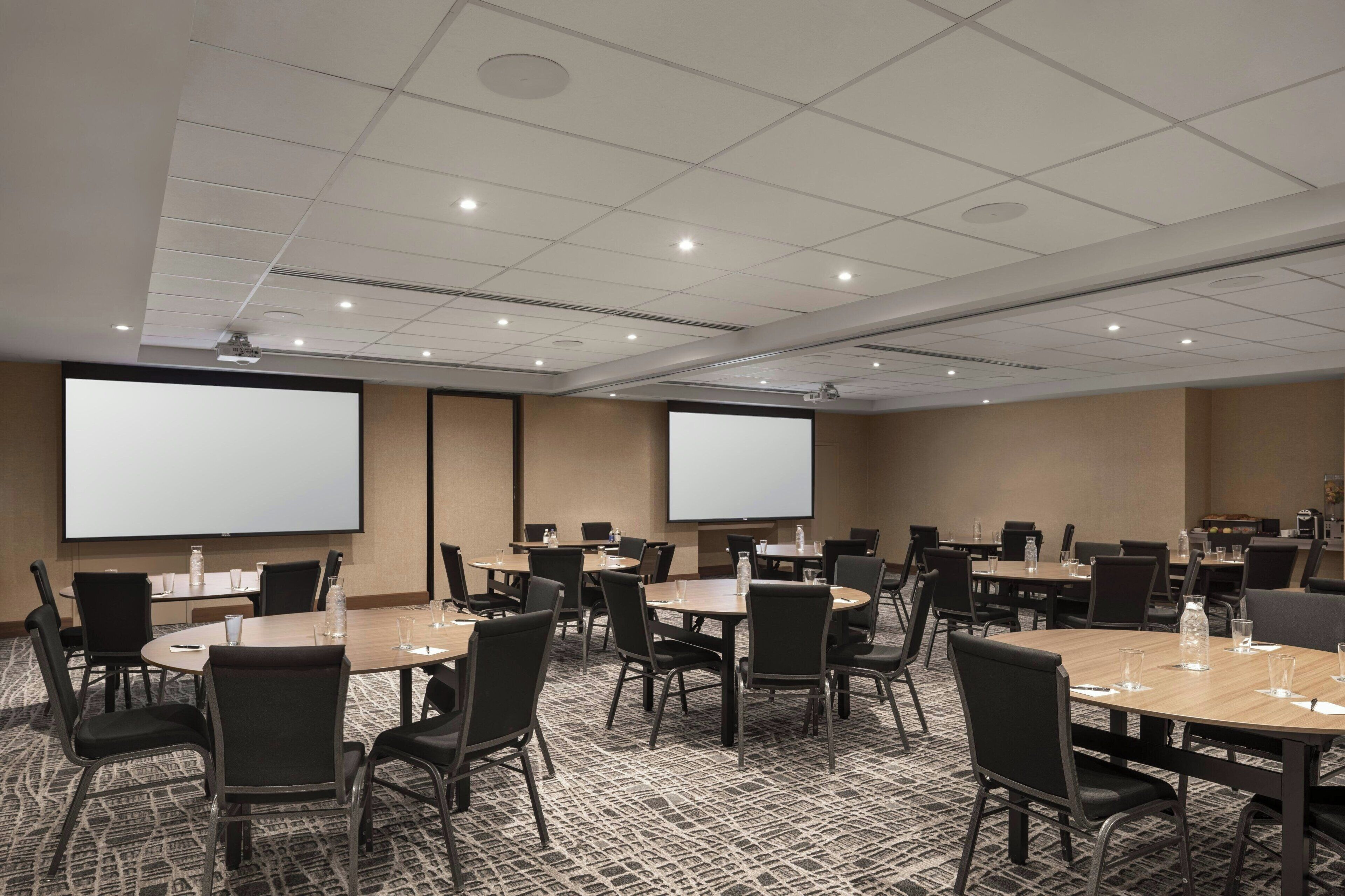 A conference room with multiple tables and chairs, a projector screen, and a carpeted floor.