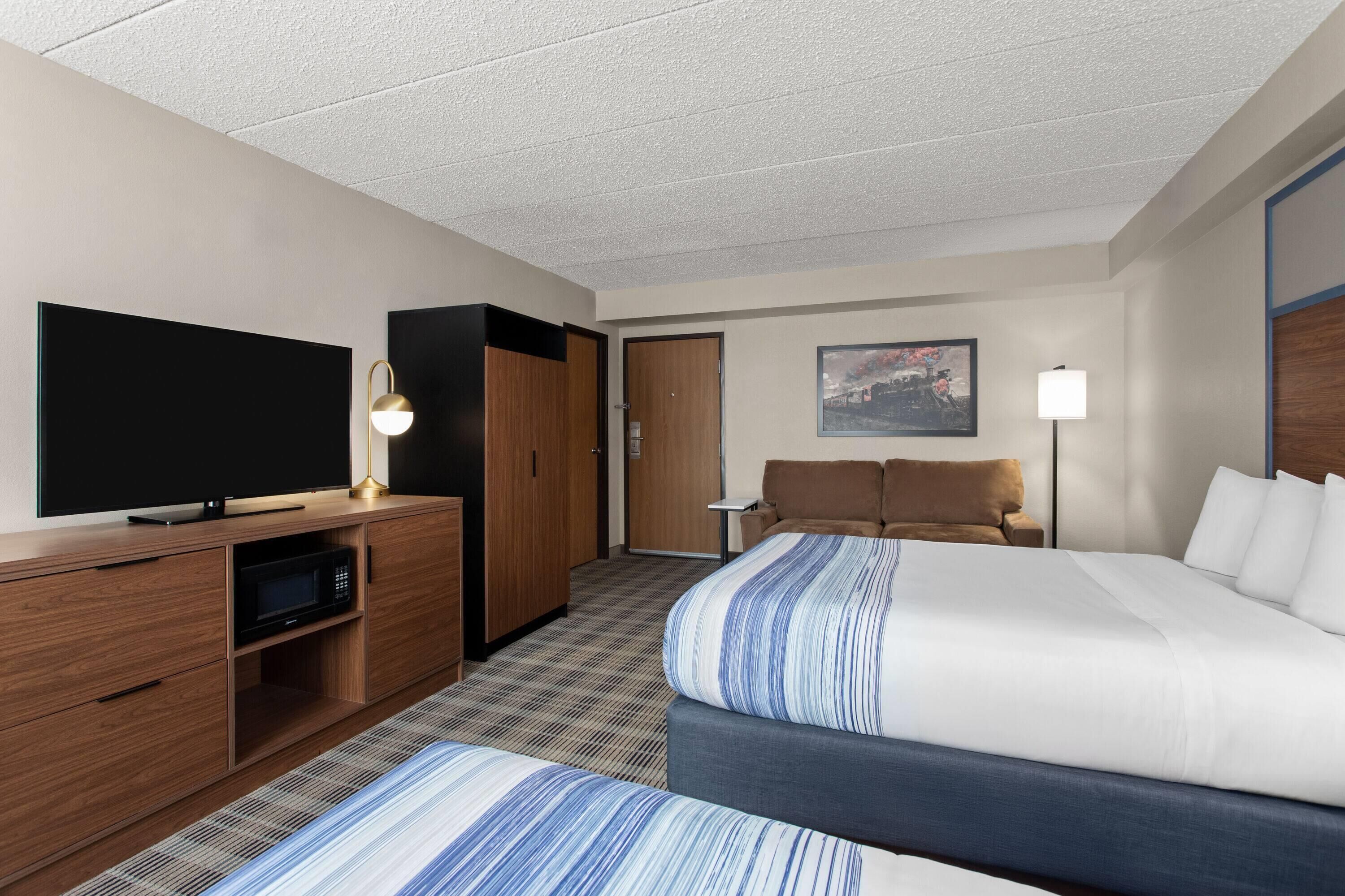 Ein Hotelzimmer mit einem großen Bett, einem Fernseher, einem Schreibtisch mit Lampe, einem Kleiderschrank und einem Bild an der Wand.