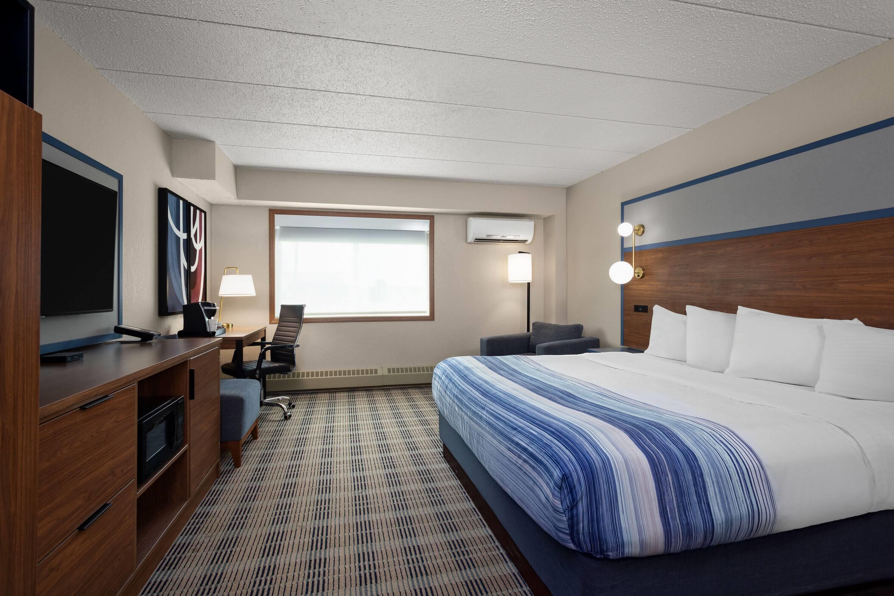 A hotel room with a large bed, a desk with a chair, a TV, and a window with natural light.