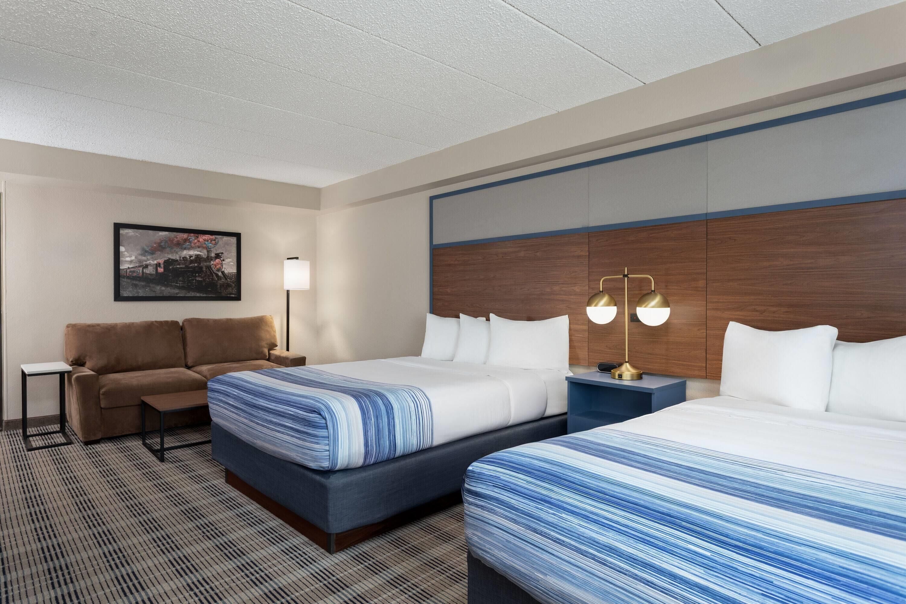 Camera d'hotel con due letti, un divano, un comodino con una lampada e un quadro appeso al muro.