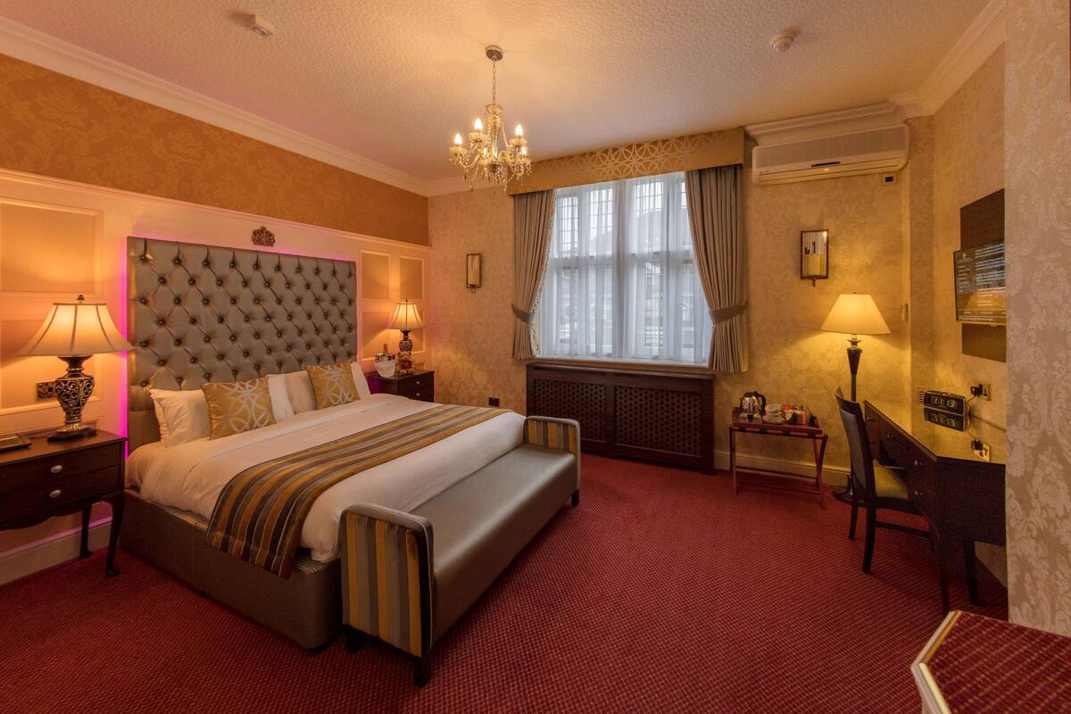 A hotel room with a large bed, two bedside tables with lamps, a desk with a chair, a radiator, and a large window with curtains.
