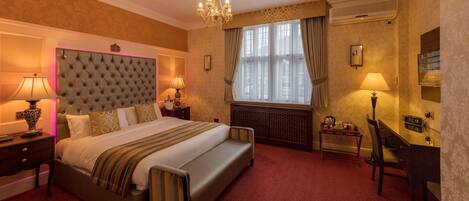 Une chambre d’hôtel avec un grand lit, deux tables de chevet avec des lampes, un bureau avec une chaise, un radiateur et une grande fenêtre avec des rideaux.