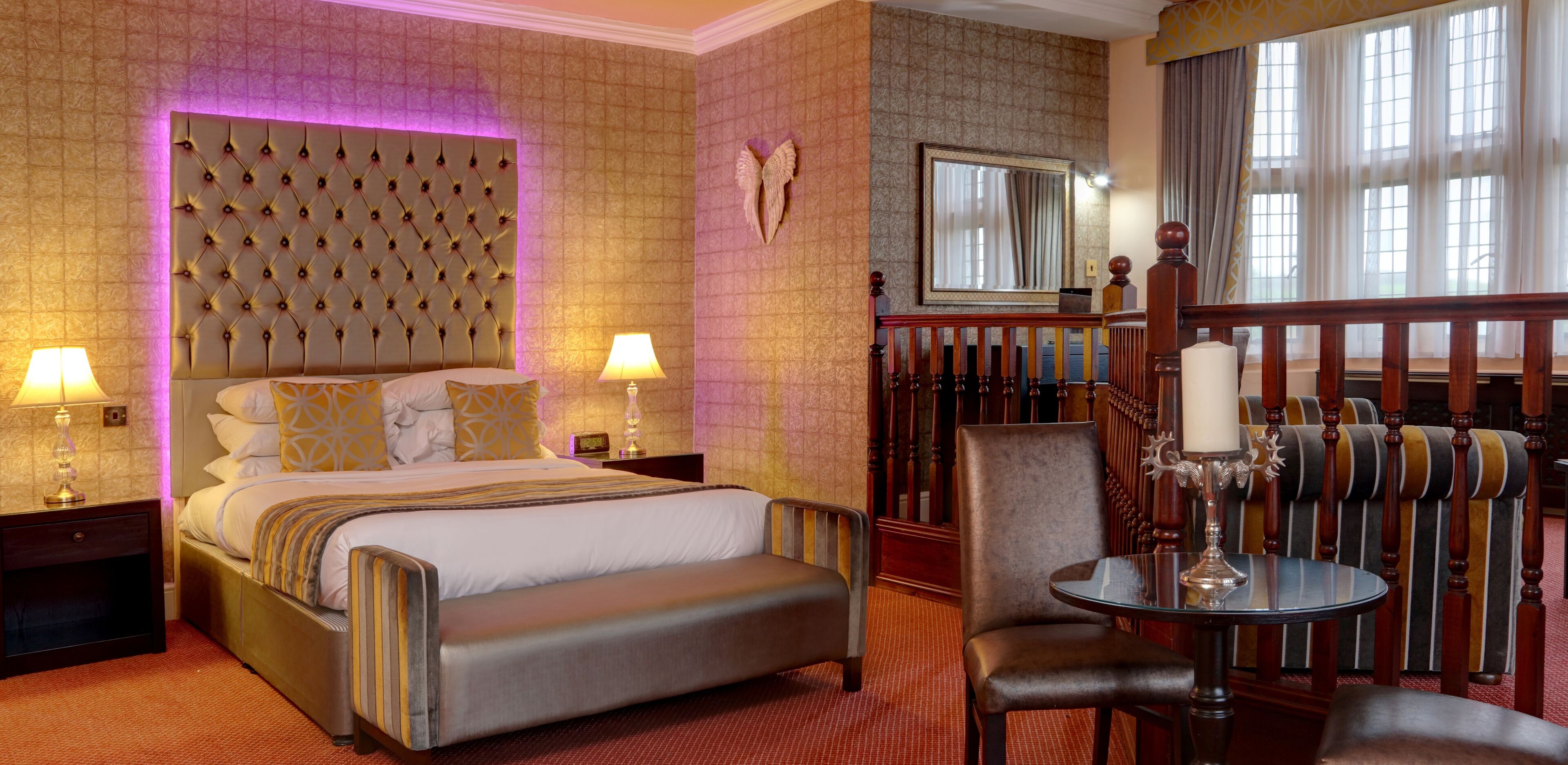 A hotel room with a tufted headboard, a bed with white linens, a bench, a small table with a candle, and a view of the outdoors through large windows.