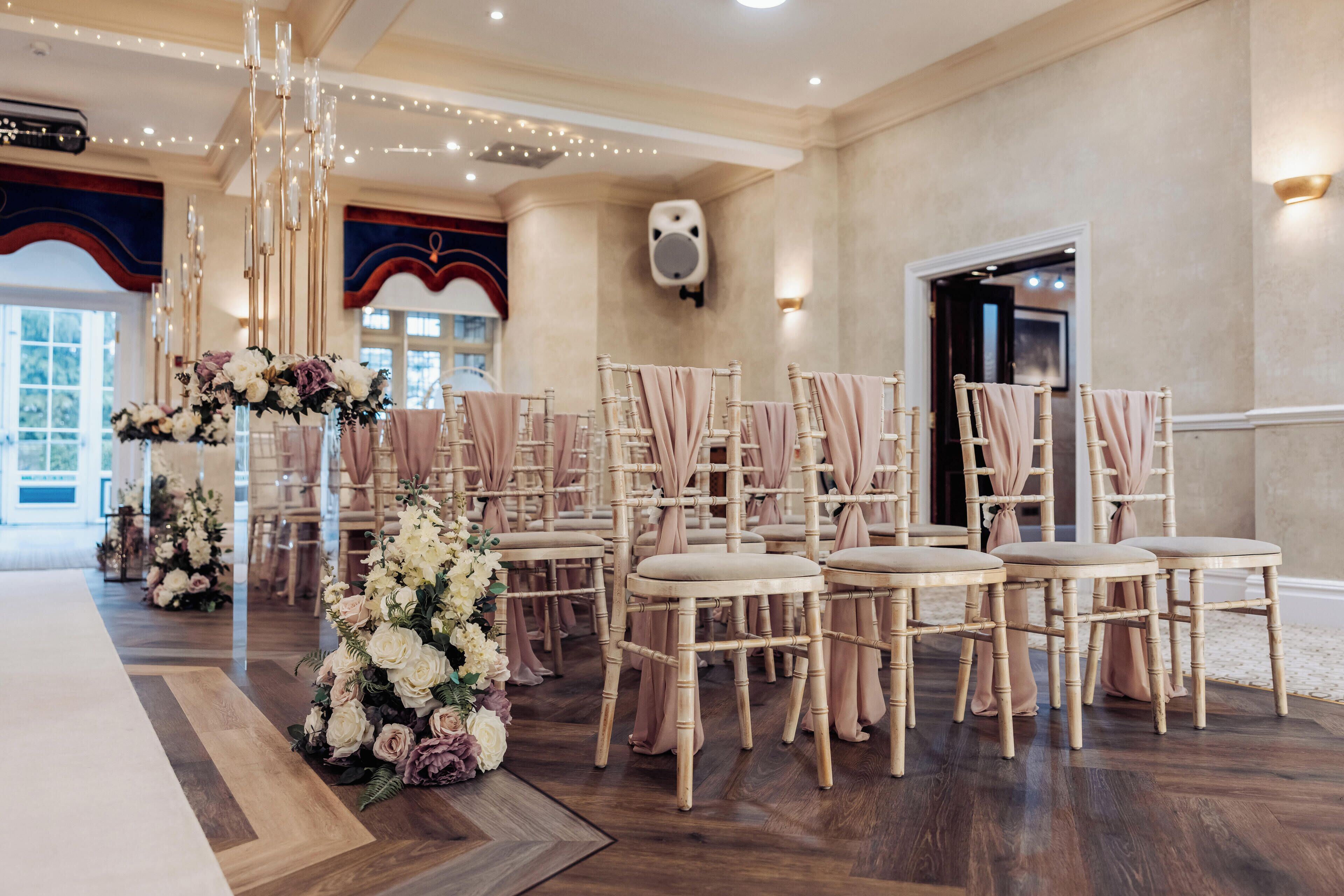 A room with wooden flooring, a floral arrangement, and rows of chairs with draped fabric.