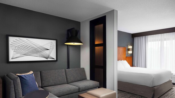 A hotel room with a gray sofa, a small wooden table, a bed with white linens, a black floor lamp, and a framed abstract artwork on the wall. - Hyatt Place Mt Laurel (Mount Laurel)
