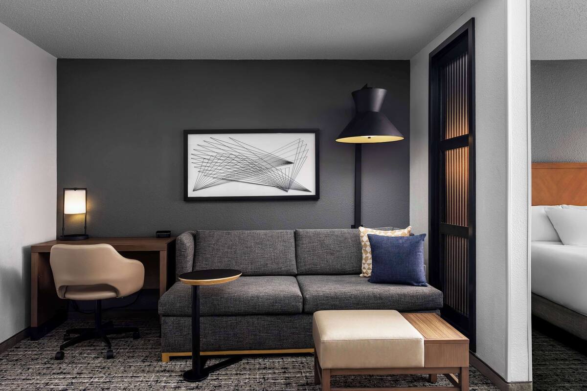 A modern hotel room with a grey sofa, a small round table, a desk with a chair, a floor lamp, and a large abstract artwork on the wall.