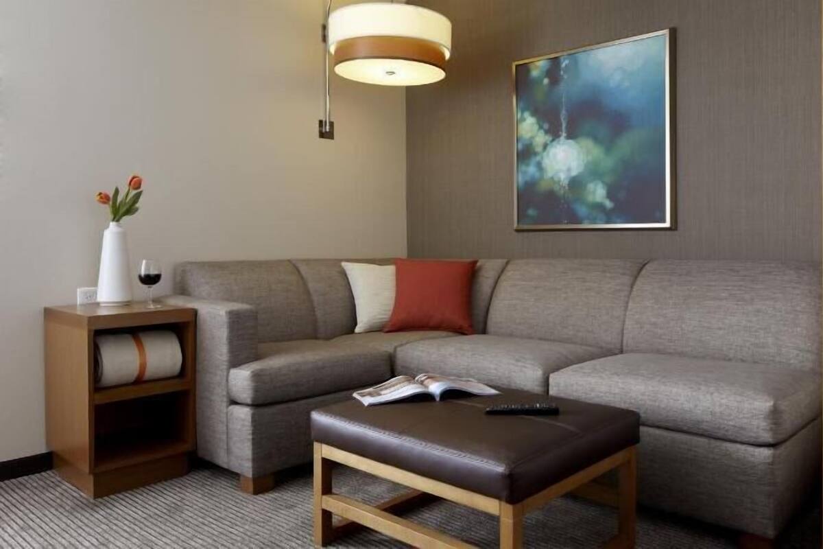 A modern living room with a grey sofa, a wooden side table, a remote control, and a book on a small wooden ottoman.