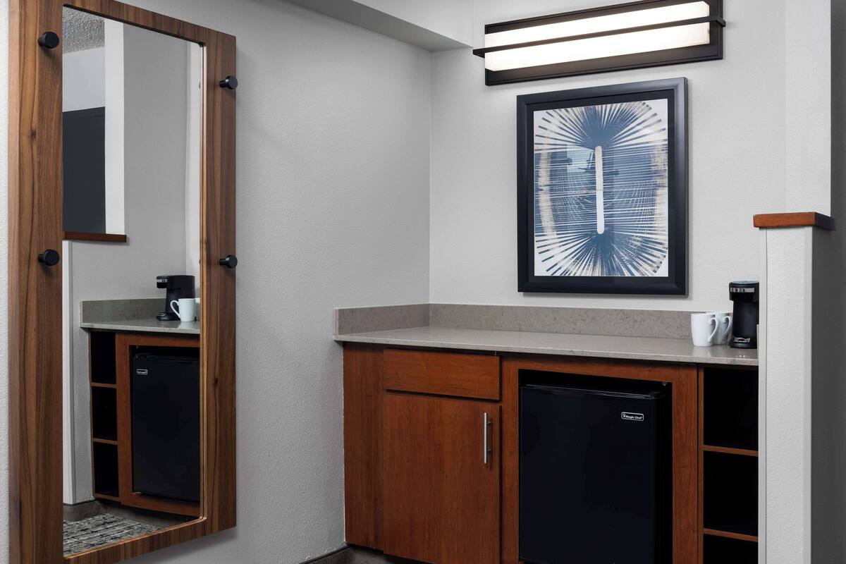 A compact bathroom with a wooden vanity, a small refrigerator, a coffee maker, and a framed abstract artwork on the wall.