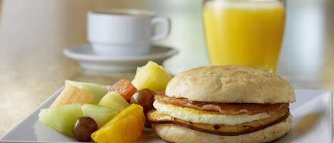Un plato de desayuno con un sándwich, fruta, una taza de café y un vaso de jugo de naranja.