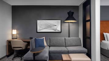 A modern hotel room with a grey sofa, a small round table, a desk with a chair, a black floor lamp, and a framed abstract artwork on the wall.