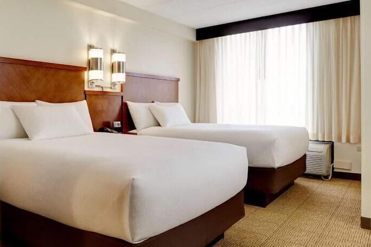 A hotel room with two beds, a wooden headboard, a nightstand, a lamp, and a window with curtains.