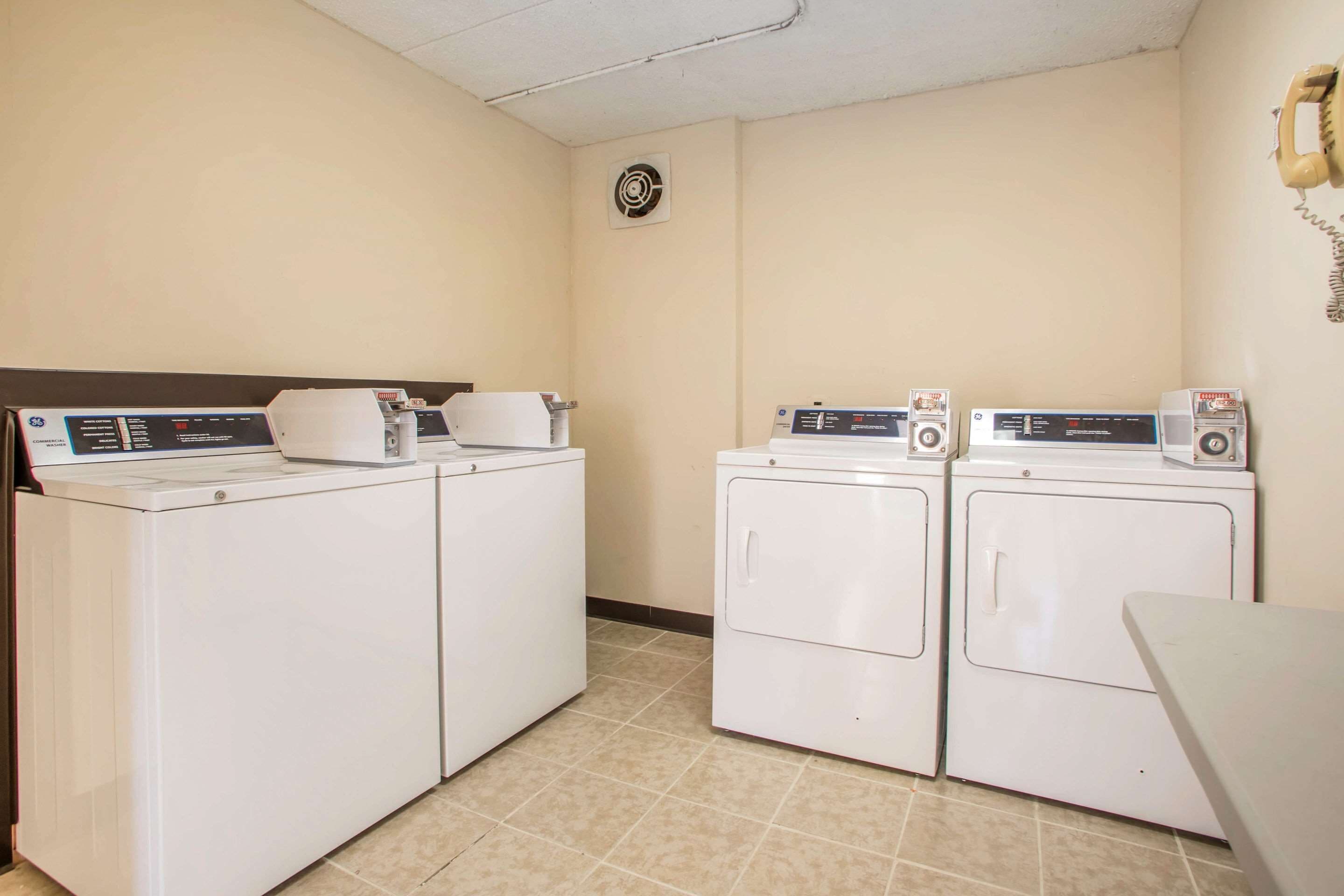 A laundry room with four white washing machines, a wall-mounted telephone, and a vent on the ceiling.