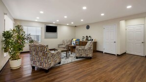 A spacious living room with a wooden floor, patterned furniture, a potted plant, and a flat-screen TV mounted on the wall.