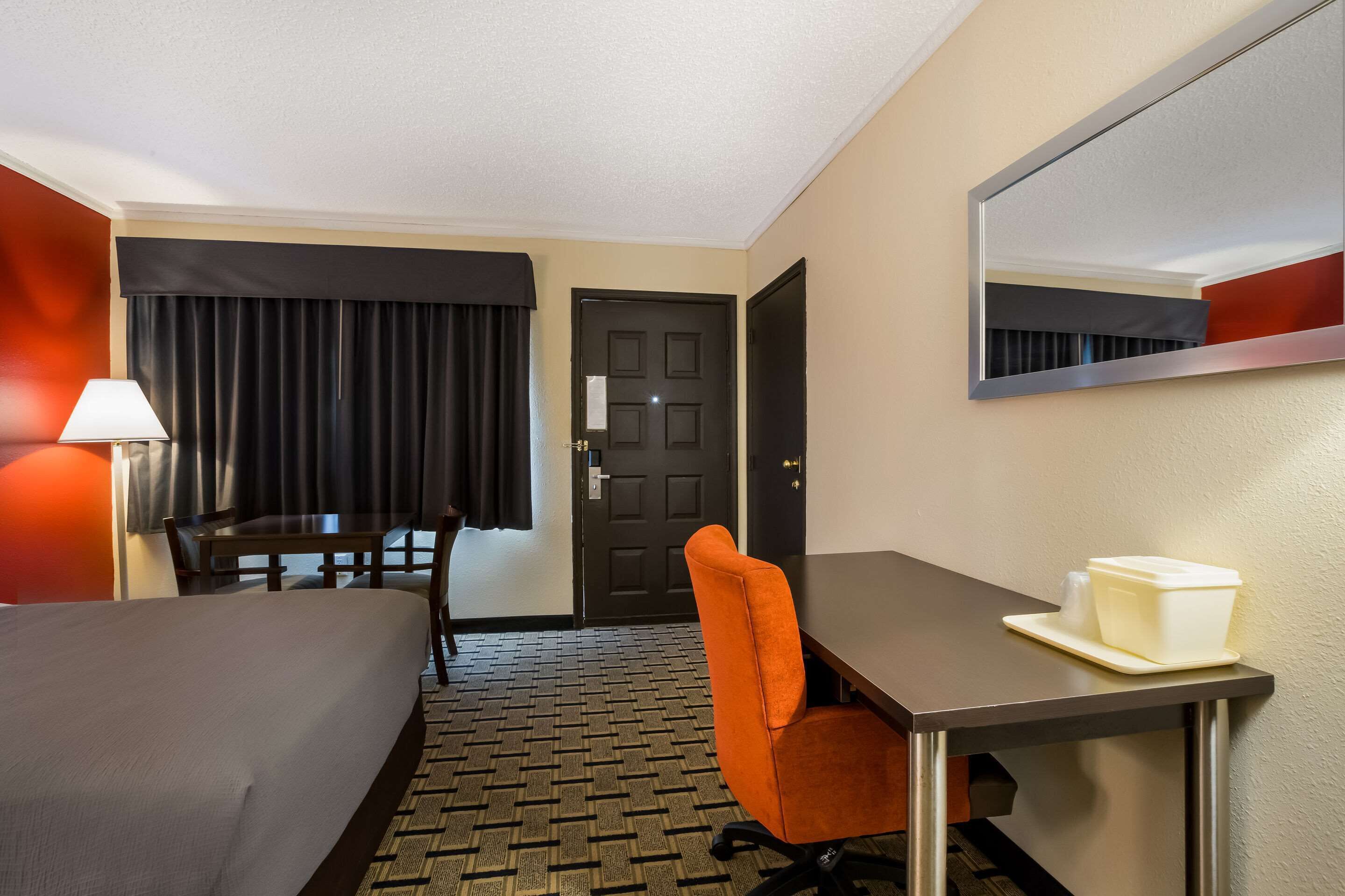 A hotel room with a bed, a desk, a chair, a lamp, a door, and a mirror.