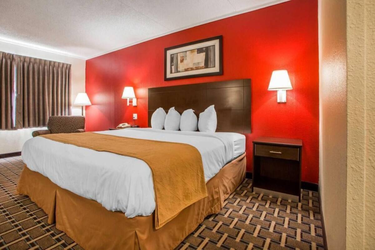 Habitación de hotel con una cama grande, dos lámparas de noche, una mesita de noche, una silla y una pared roja con un cuadro enmarcado.