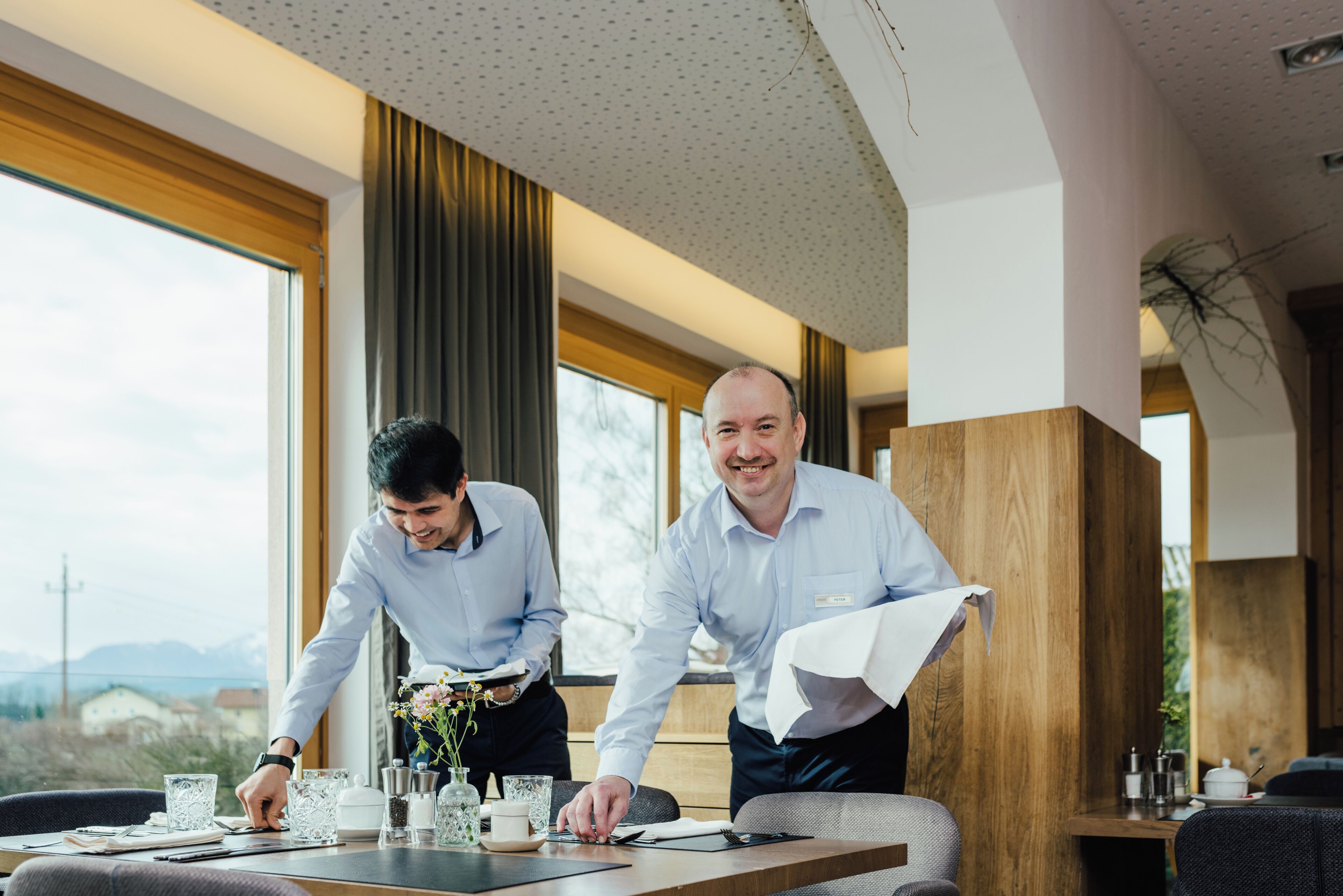 Twee mannen in een moderne eetruimte met grote ramen, een houten accentmuur en uitzicht op bergen.