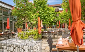 An outdoor dining area with tables and chairs, potted plants, and orange umbrellas. - Hotel Ammerhauser (Anthering)