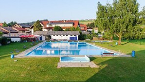 Une piscine extérieure rectangulaire, dotée d’un plongeoir, entourée d’une pelouse et avec un bâtiment au toit rouge en arrière-plan.