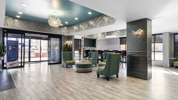 A spacious lobby with a modern design, featuring a large seating area with green upholstered chairs and a central ottoman, a large flat-screen TV mounted on a textured wall, and a patterned ceiling with recessed lighting. - Quality Inn & Suites Montgomery East Carmichael Rd (Montgomery)