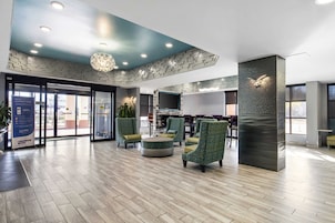 Un vestíbulo espacioso con diseño moderno, que cuenta con una amplia zona de estar con sillones tapizados en verde y un otomán central, un televisor de pantalla plana grande montado en una pared texturizada y un techo con diseño con iluminación empotrada.