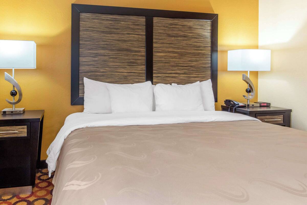 A hotel room with a large bed, two bedside tables, a nightstand, and a wall-mounted lamp.