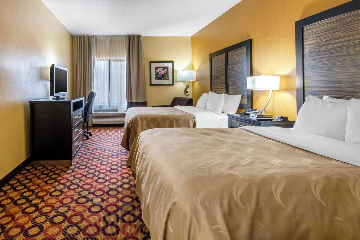A hotel room with two beds, a desk with a computer, a chair, a lamp, and a window with curtains.