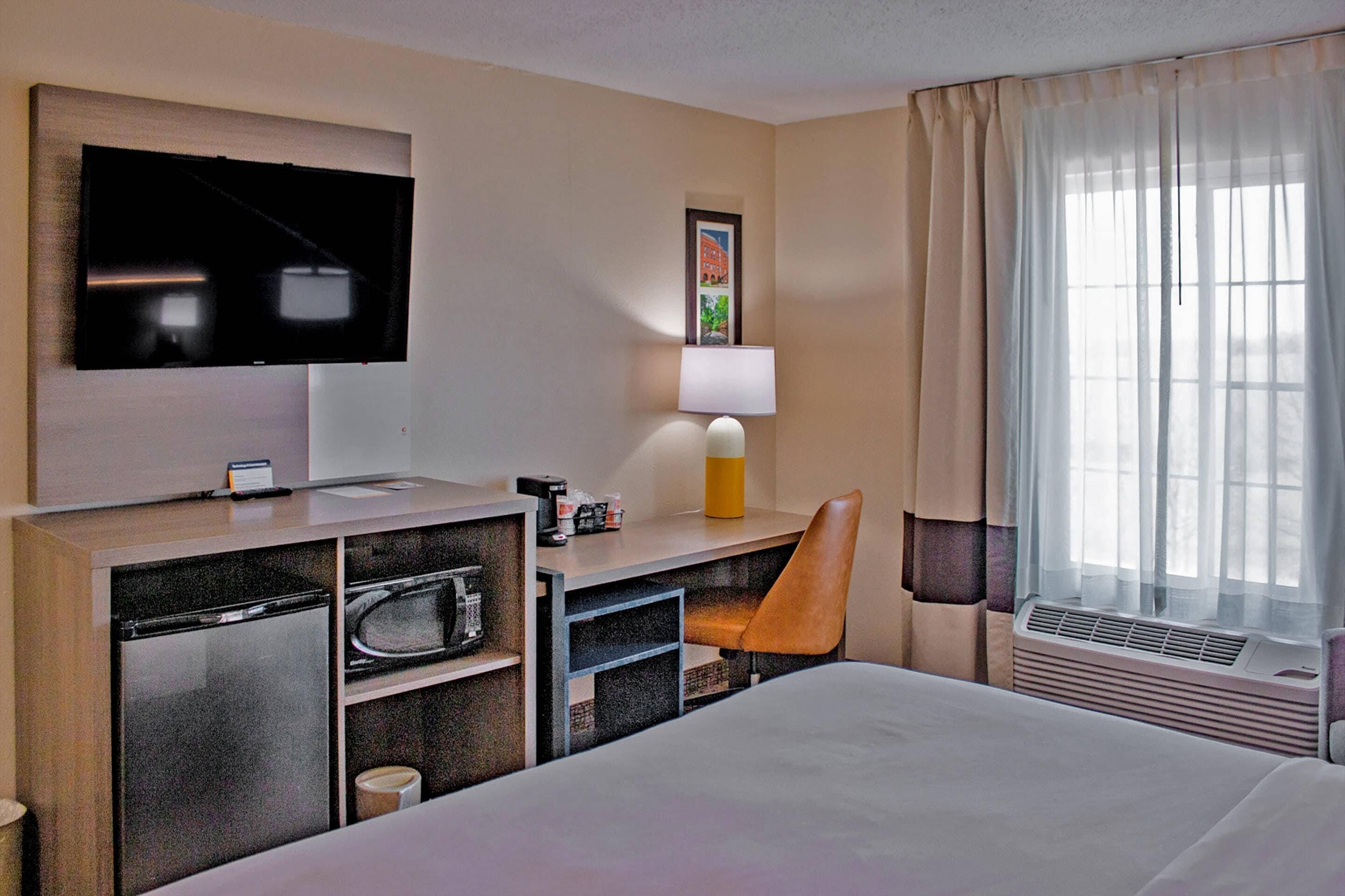 A hotel room with a bed, a desk with a television, a chair, a microwave, a mini-fridge, a lamp, and a window with curtains.