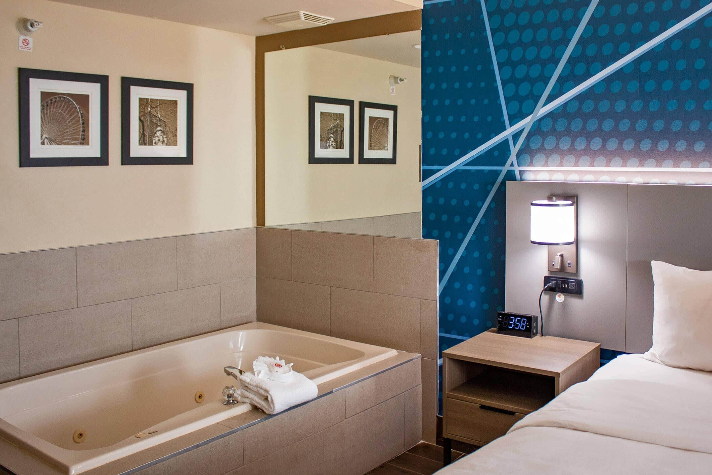 A hotel room with a large bathtub, a bed, a nightstand with a clock, and framed pictures on the wall.