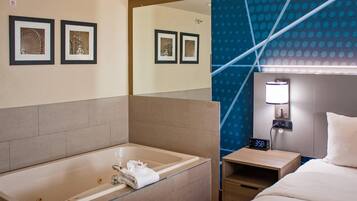 A hotel room with a large bathtub, a bed, a nightstand with a clock, and framed pictures on the wall.