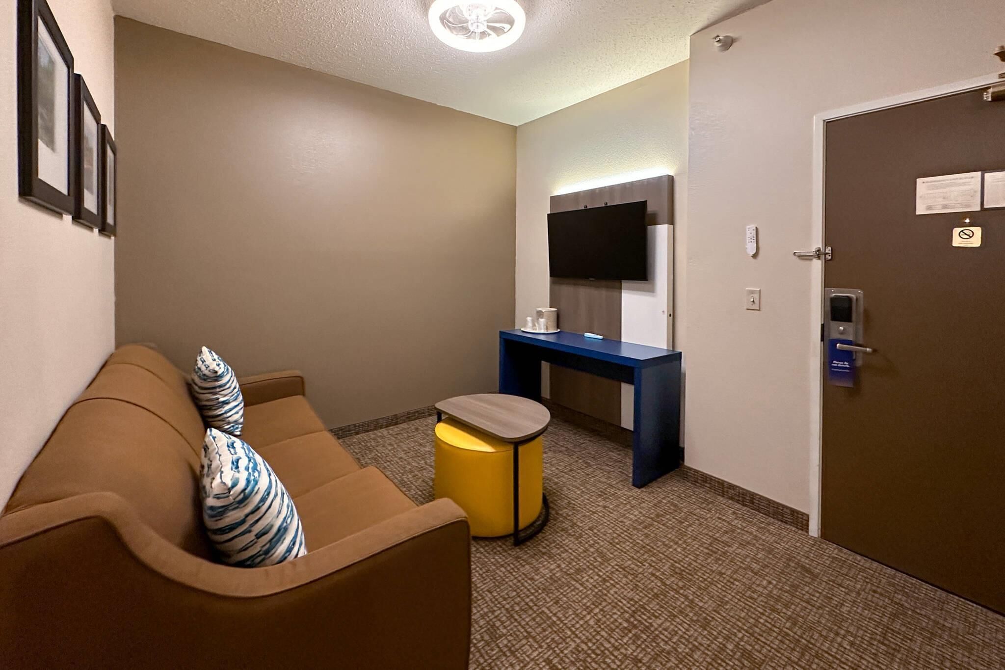Habitación de hotel con un sofá marrón, un pequeño taburete amarillo, un escritorio azul y un televisor montado en la pared.