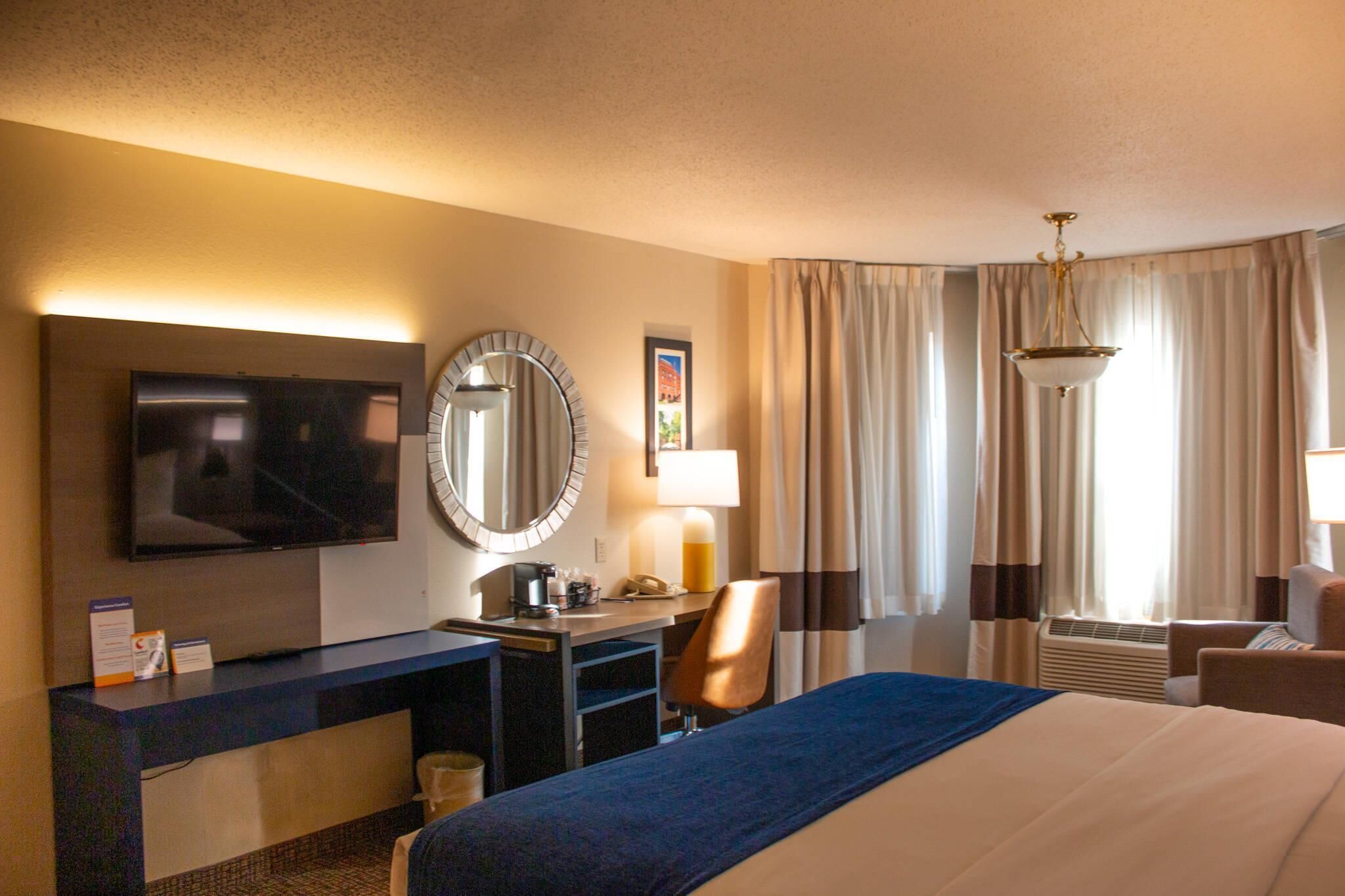 A hotel room with a bed, desk, TV, and a large mirror.