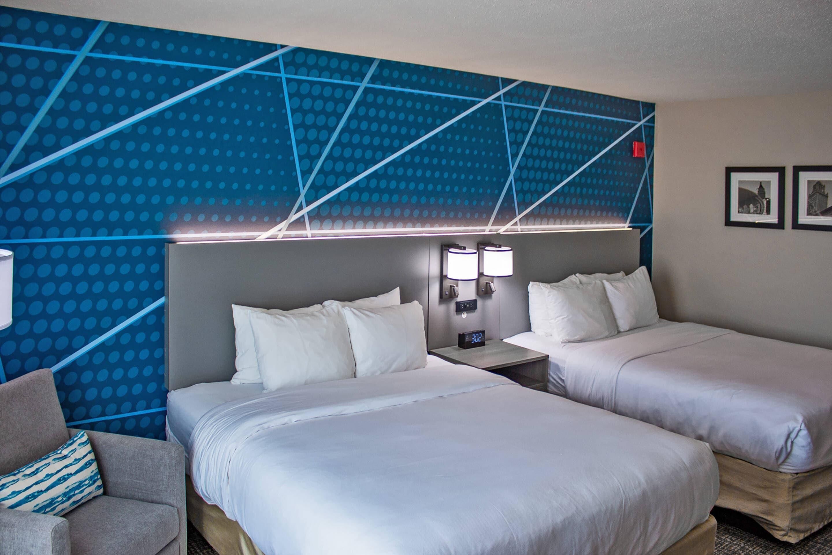 Habitación de hotel con dos camas, una mesita de noche con un reloj y una pared con diseño geométrico.