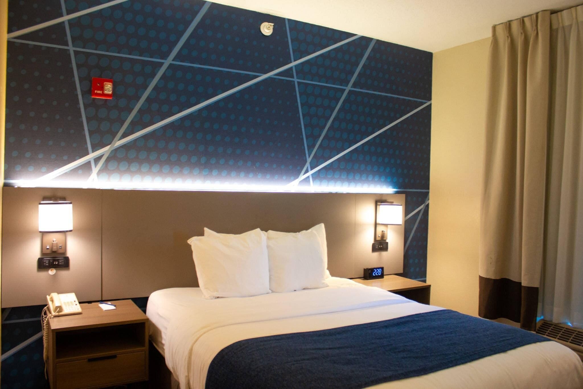 A hotel room with a large bed, two bedside tables, a nightstand, a wall-mounted phone, and a digital clock.
