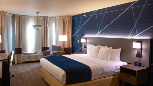 Ein Hotelzimmer mit einem großen Bett, zwei Nachttischen, einem Schreibtisch und einem Stuhl. Es gibt ein Fenster mit Vorhängen, eine Wandleuchte und eine Deckenlampe.